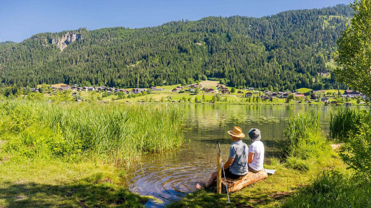 Sommeridylle am Weissensee: Zwei Menschen, einer davon mit Blindenstock, sitzen am Ufer und kühlen ihre Füße im Wasser