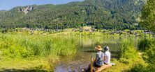 Sommeridylle am Weissensee: Zwei Menschen, einer davon mit Blindenstock, sitzen am Ufer und kühlen ihre Füße im Wasser