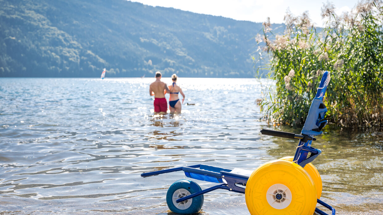 Ein Paar geht im Millstättersee schwimmen, im Vordergrund ein Strandrollstuhl